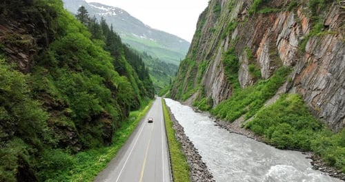 A car approaches by the road built along the fast narrow river at the mountain foot.