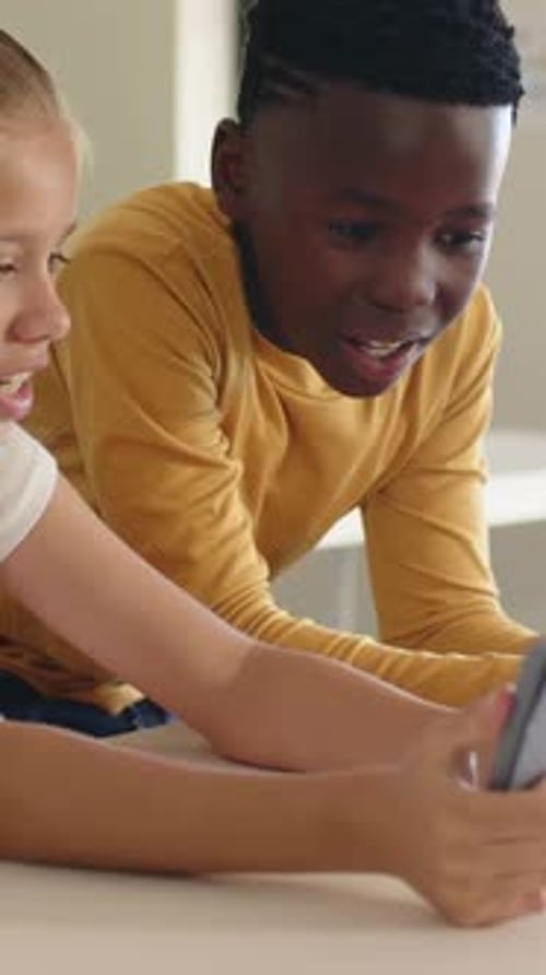 Vertical video: Leaning yellow-shirted boy and light-topped girl watching tablet clip on white desk