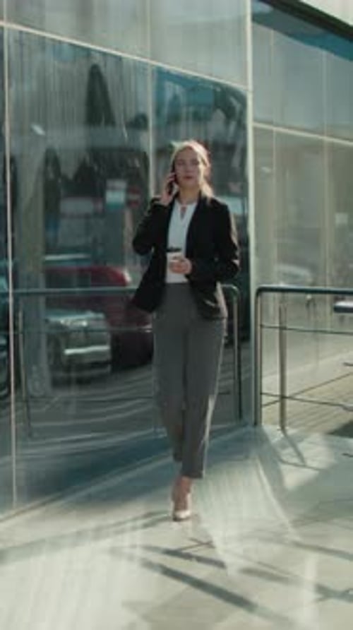 Elegant Lady Walking with Coffee on Phone Reflected on Glass Building Exterior
