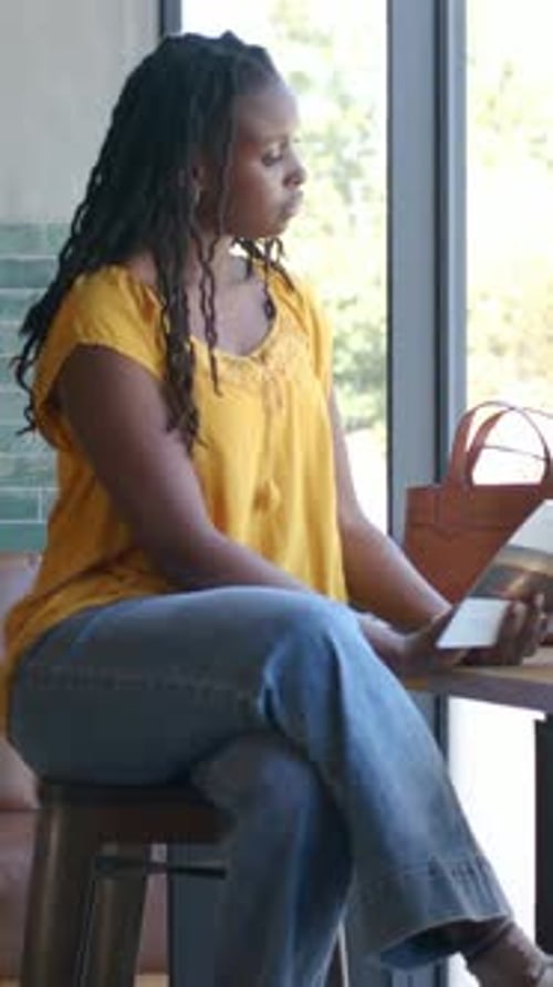 Vertical video: Holding card, woman in yellow top flipping it at cafe window near brown bag waiting