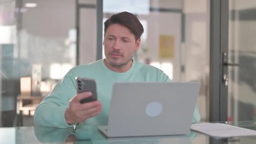 Man Uses Phone and Laptop in Bright Office