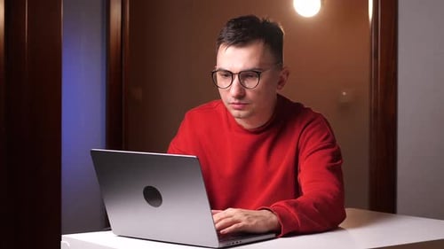 Gestresster junger Mann mit Brille aufgrund von Computerüberlastung, sitzender Arbeitsalltag