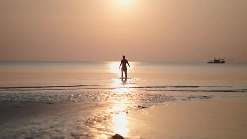 Young Man Walking Towards the Rising Sun Reflected in the Shallow Waters of a Tropical Beach
