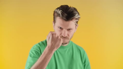 A Man with Stubble Stands Against a Yellow Wall and Shakes His Fist at the Camera A Man in a Green