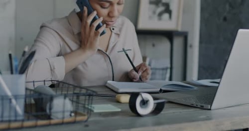 Female Office Worker Speaking on Mobile Phone and Taking Notes Working at Desk Alone
