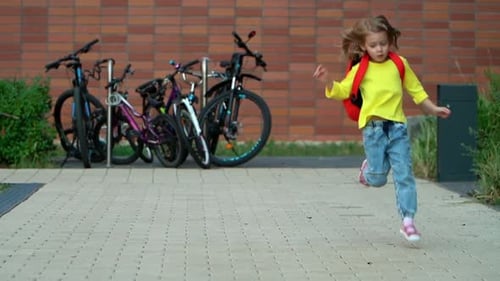 Little Happy Student Girl Runs and Jumps with Backpack in School Yard Education