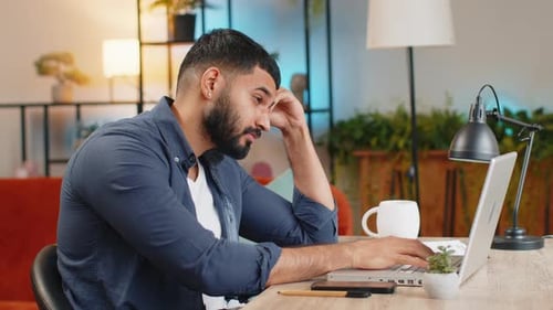 Bored Sleepy Young Indian Man Working on Laptop Computer Yawning Leaning on Hand at Home Office