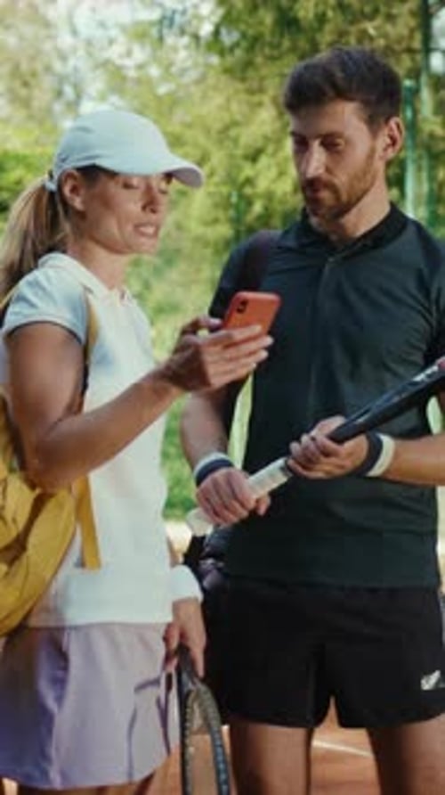A Man and Woman Stand on a Tennis Court Looking at a Phone