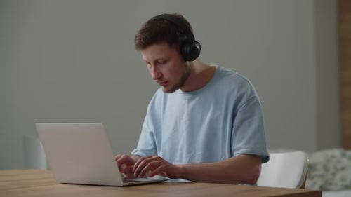 View of a Young Man in a Blue Tshirt Who Listens to Music While Working on a Laptop A Male Student