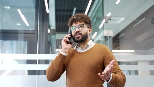 Confident businessman talking on mobile phone while standing in modern glass office.