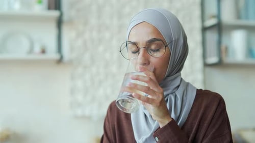 Muslim woman freelancer drinks clean water while sitting at table at home Happy female with a glass