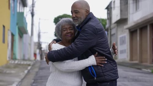 Caring African American adult son in 50s hugging his elderly mother outside in urban street