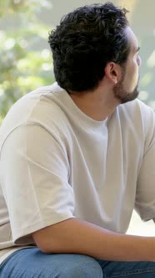 Vidéo verticale : Tourner un homme sur un siège lumineux en t-shirt beige sur le porche, la caméra bouge, ferme les yeux, sourit