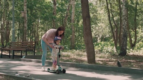 Mom Teaches Daughter to Ride a Scooter in the Park in Summer