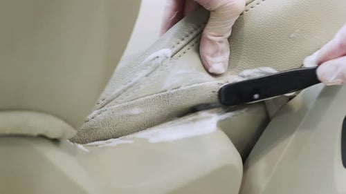 Detailing of the interior of a modern car. A man cleans a beige car seat with a brush and cleaning s