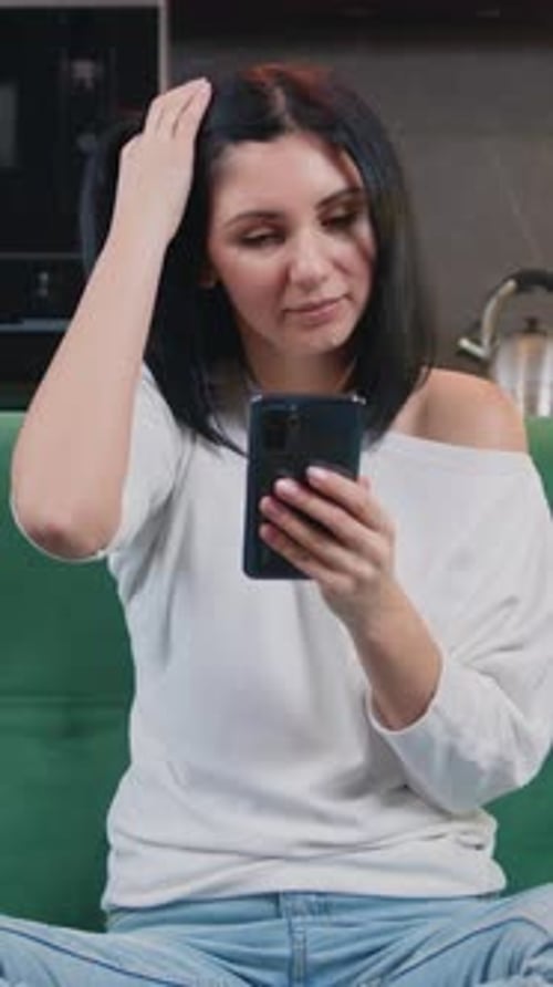 Happy Young Woman Sitting on a Green Sofa and Using a Smartphone Smiling in a Modern Kitchen Female