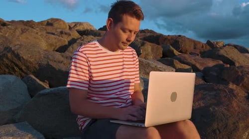 Young Caucasian Man Remotely Works with Laptop on Rocky Beach at Sunset Guy is Sitting on Rocks with