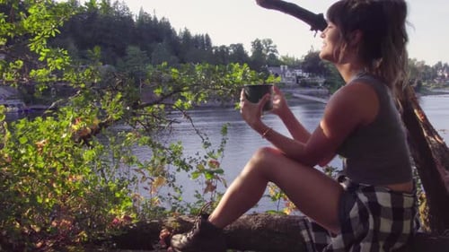 Cute hipster girl sitting by a lake with a mug to drink and relax