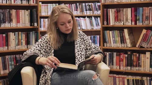 Female is sitting on chair and reading a book in library, dolly shot
