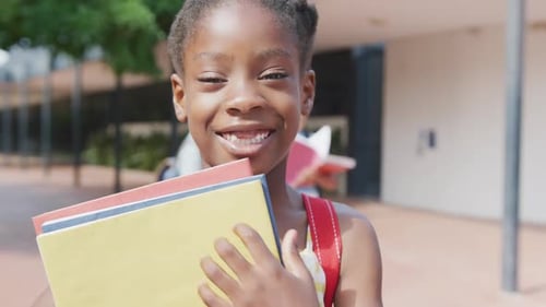 Video portrait of happy african american schoolgirl holding her books outside school