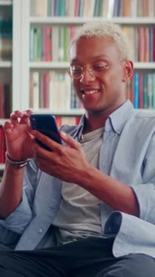 Young Adult Uses Phone at Home by Bookshelf