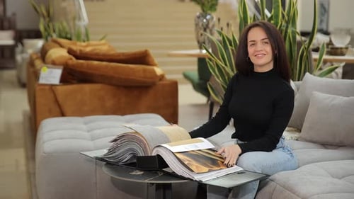 Smiling Woman Choosing Furniture and Reading Catalog on Sofa in Store