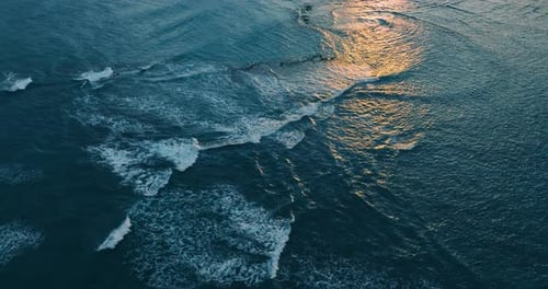 Foamy waves roll by the blue waterscape. Setting sun reflects in the ocean.