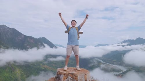 Homem de pé na rocha e levantando as mãos comemorando a chegada ao topo da montanha nebulosa