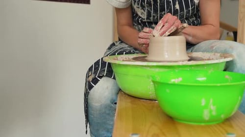A female potter works on a potter's wheel, making a ceramic pot from clay in a pottery workshop.