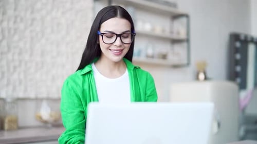 Smiling brunette with eyeglasses using laptop in the light room. Attractive happy woman in green