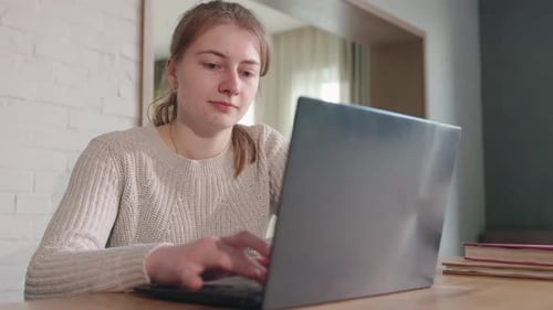 Young Woman Sits at a Table and Works on a Laptop with Focused Expression During Online Learning or