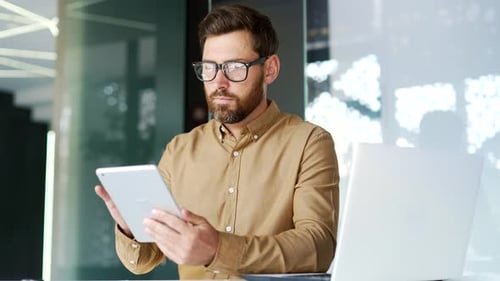 Manager is using digital tablet while sitting at workplace in business office. Worker reads