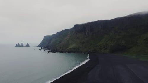 Aerial drone shot of beautiful blue waves crashing on a black sand beach in Iceland and huge rock fo