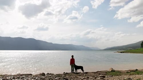 Girl Stands Near Dog of Rottweiler Breed on Shore Near Lake Against Backdrop of Mountain Range