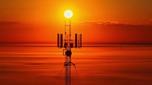 Cell tower with bright sunlight glowing behind it, filmed by drone
