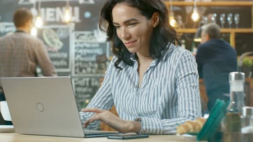 Attractive Young Woman Drinks Beverage and Works on a Laptop Computer while Sitting in the Stylish