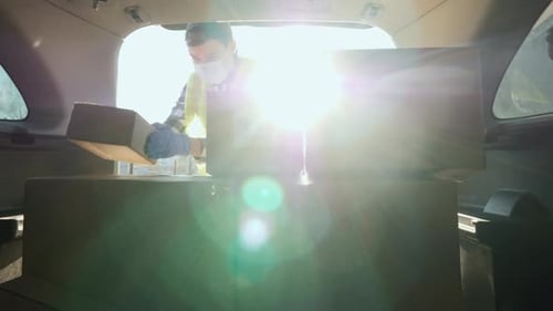 A Young Man in a Medical Mask Loads Boxes of Medicines in the Trunk of a Car