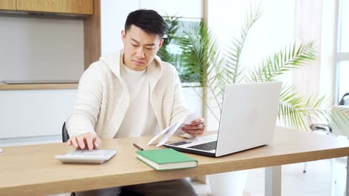 young Asian man counts bills, bank checks, loans or utilities while sitting at home at a kitchen