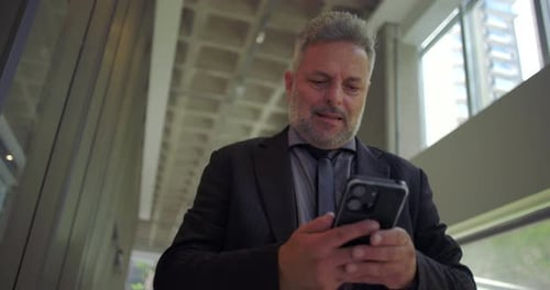 Middle aged businessman standing using smartphone, slightly smiling while checking screen