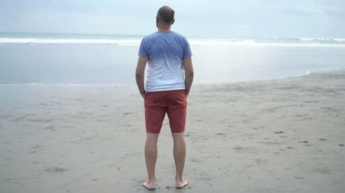 Junger Mann steht gedankenverloren am Sandstrand und bewundert den weiten Meerblick