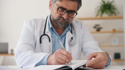 Writing information into notepad. Mature male doctor in coat is in the clinic room
