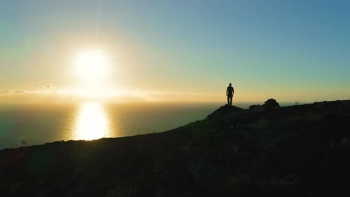 Man Silhouette Stands on Edge of Cliff and Looks Out at Ocean During Sunset