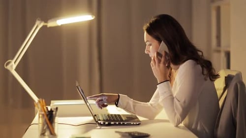 Businesswoman calls on smartphone while working late with laptop in night office