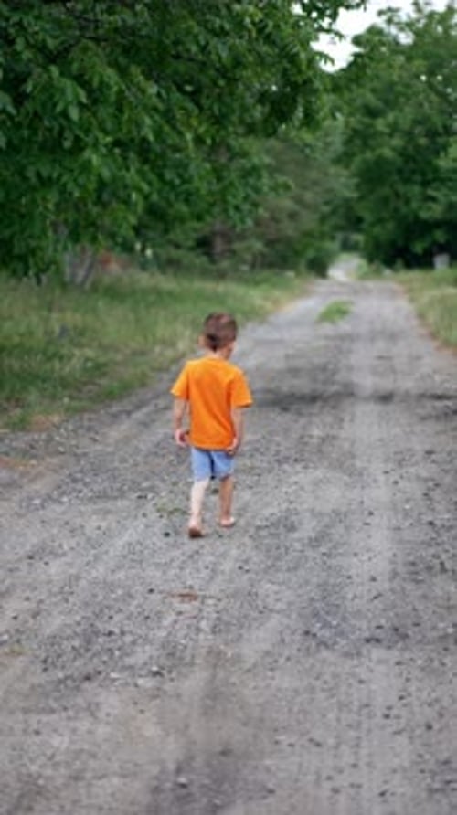 Ein kleines barfüßiges Kind in orangefarbenem T-Shirt und blauen Shorts rennt die Landstraße entlang.