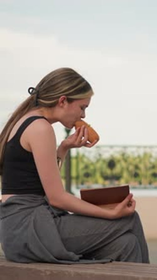 Woman Eating Burger and Reading Outdoors Near Decorative Fence Under Bright Sky