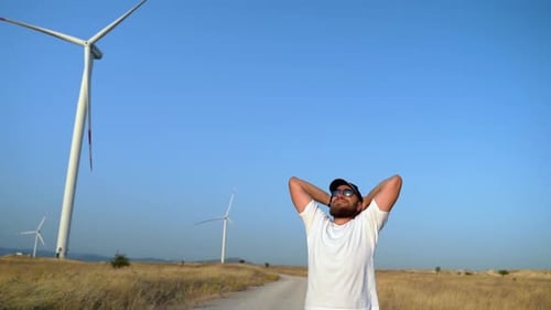Feliz hombre barbudo relajado levantando los brazos disfrutando de un viaje en la naturaleza Puesta de sol en el campo molino de viento