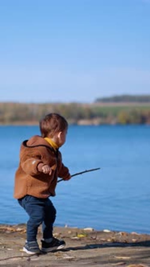 Little Caucasian kid is on the river bank. Baby boy puts the stick into water.
