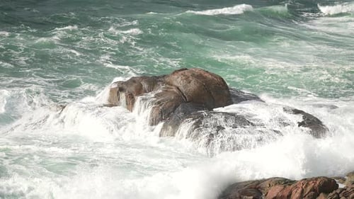 Powerful ocean waves crash over large rocks at a rugged shoreline, with white foam splashing