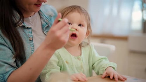 Mother Feeding Blonde Toddler with Spoon in Home