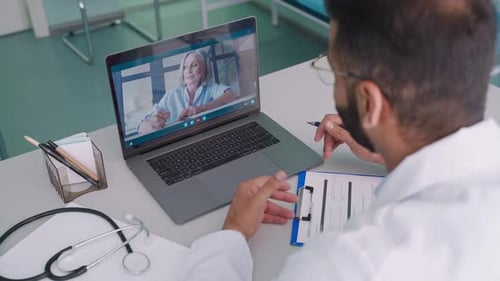 Videocall Chat With Female Older Patient And Indian Male Doctor In Modern Clinic Hospital Using P...